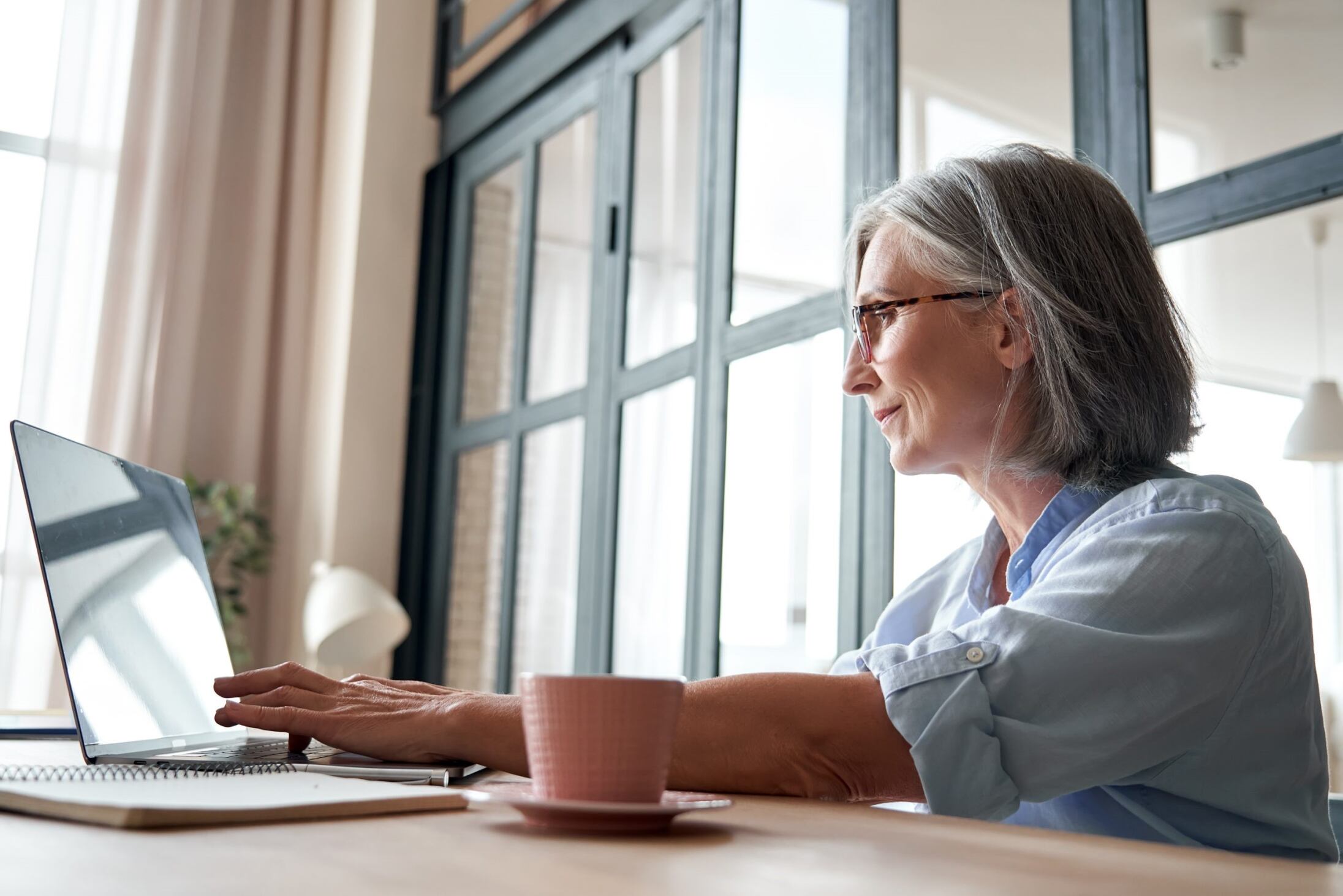 Senior professional woman with silver hair working at laptop by window, representing workplace menopause inclusion
